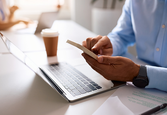 Online Payroll Services for Small Businesses - PEOPayGo - PEOPayGo Close-up of hands holding a smartphone over an open laptop on a clean office desk with a coffee cup nearby?