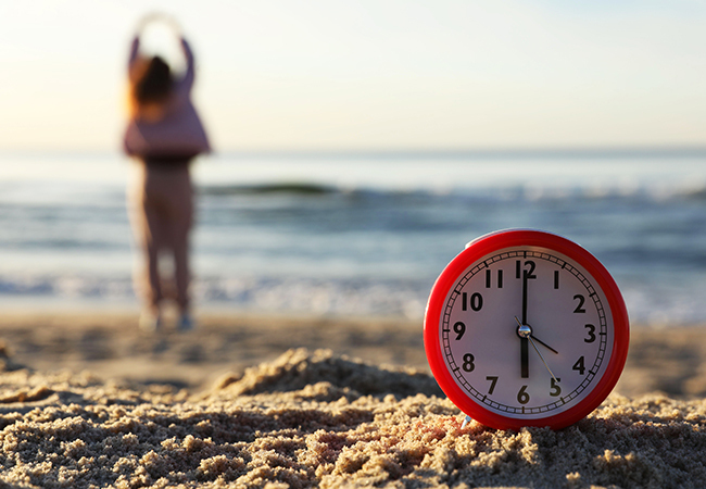 PTO vs Vacation Time Which Policy is Better_ - PEOPayGo - PEOPayGo Red alarm clock on the sandy beach with the ocean in the background and a blurred person stretching nearby beyond the clock.