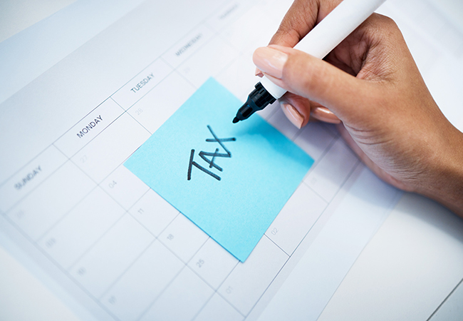Payroll Tax Compliance Rules Requirements - PEOPayGo - PEOPayGo Hand writing 'Tax' on a blue sticky note placed on a calendar, signaling tax deadline reminder.