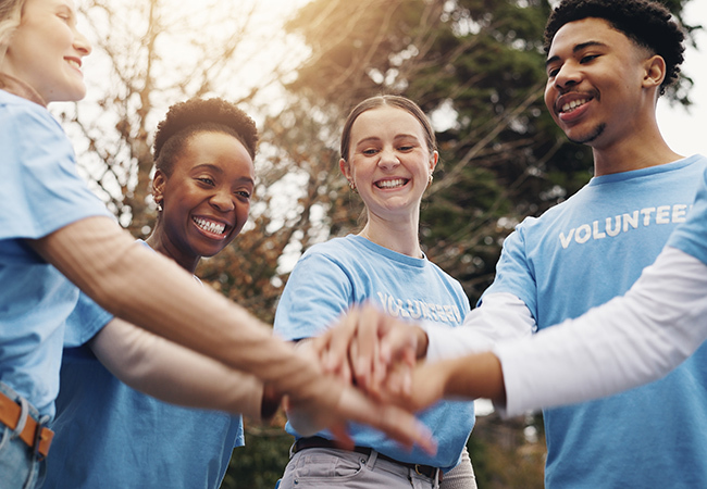 Nómina para organizaciones sin fines de lucro Reglas Requisitos fiscales - PEOPayGo Group of diverse volunteers in blue shirts join hands in a circle outdoors, smiling and showing teamwork.