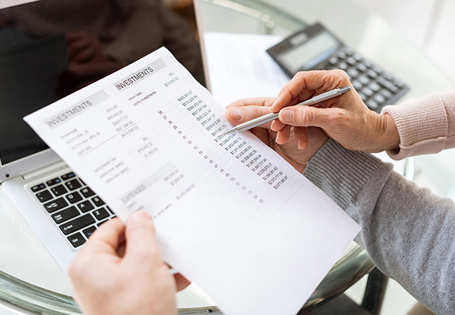 Person reviewing an investment statement with a pen, a laptop nearby on a glass table and a calculator in view.