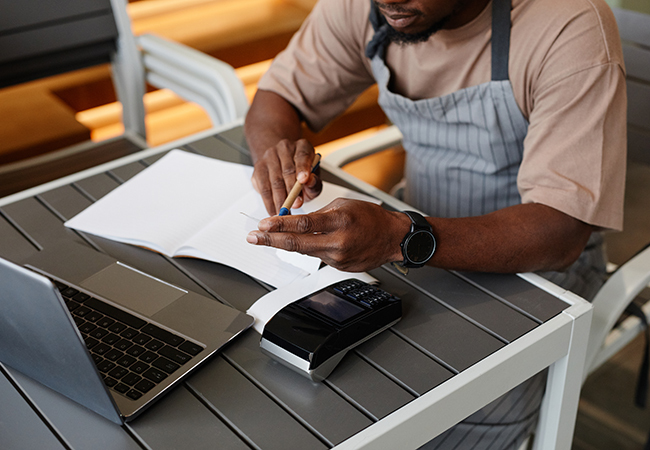 What Are the Key Payroll Process Steps for Small Business_ - PEOPayGo - PEOPayGo Man in apron writes in a notebook at an outdoor table, with a laptop and card reader beside him.