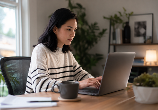 Workers comp for remote employees - PEOPayGo - PEOPayGo Woman in a striped sweater typing on a laptop at a wooden desk in a home office.