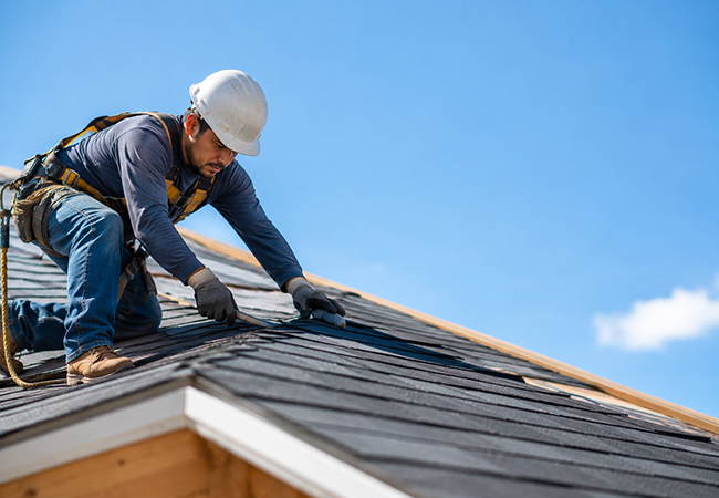 Workers comp for roofers - PEOPayGo - PEOPayGo Construction worker on a sloped roof, installing dark shingles while wearing a hard hat and safety harness.